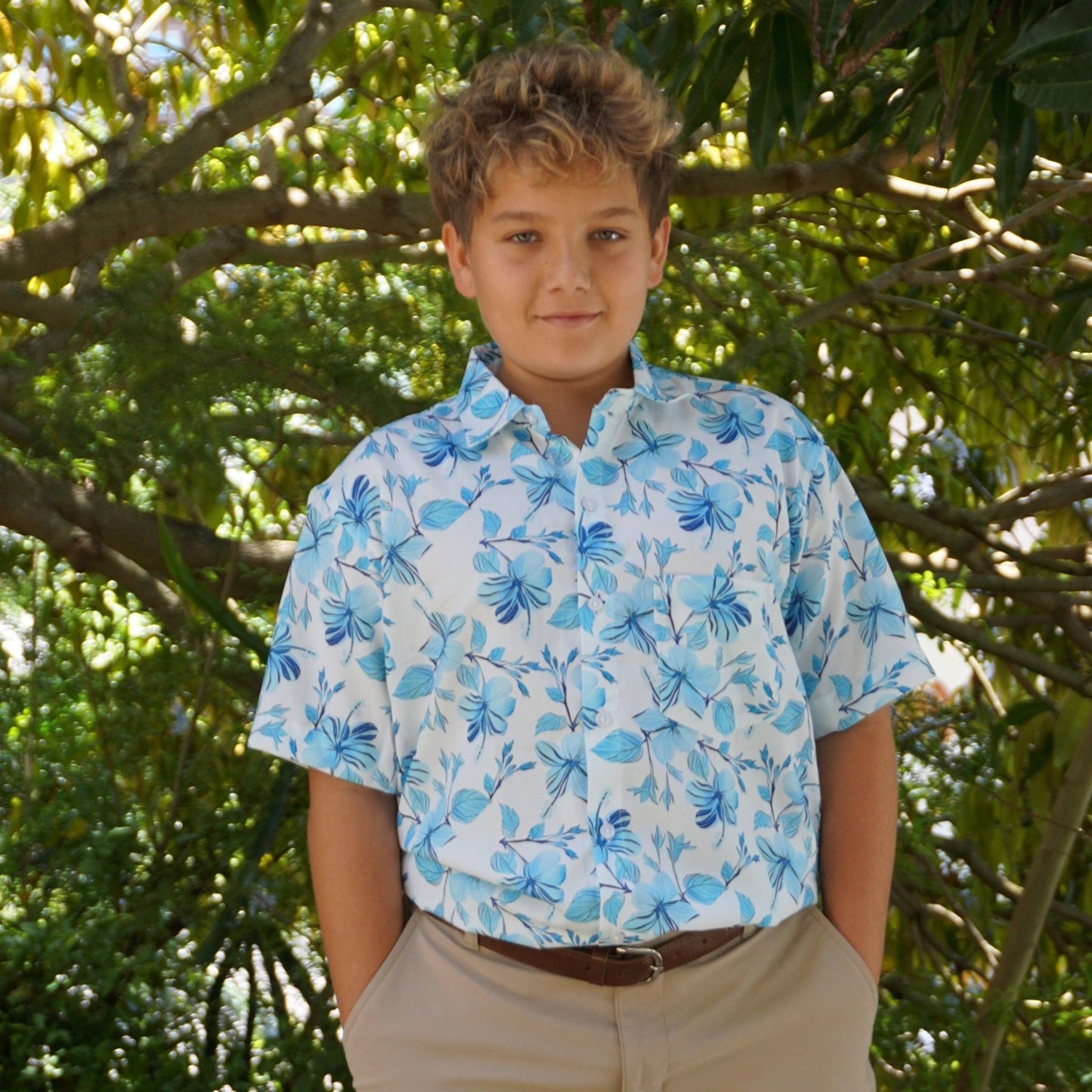 A white shirt with a light blue hibiscus flower pattern, an aloha Hawaiian Shirt, on a young model in a lush forest. Made by Phin Hawaii