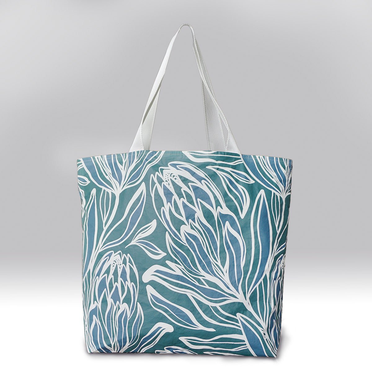 The back view of a blue and green colored tote bag with a white protea flower design. Has two pockets inside, with a zipper closure.